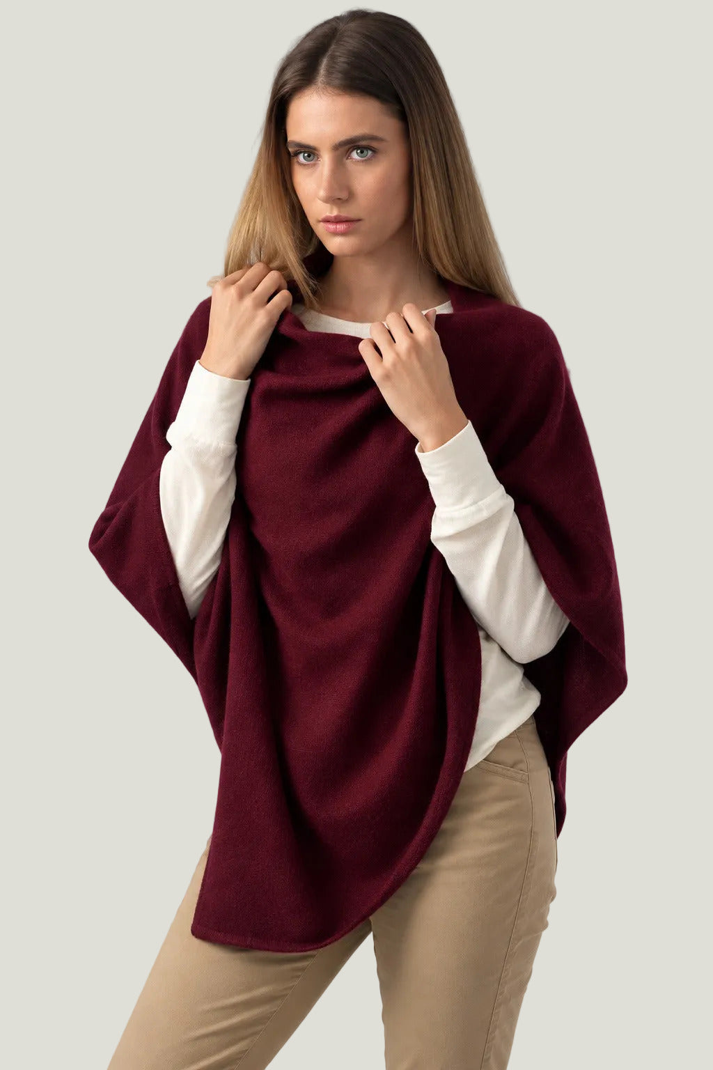 A woman modeling a deep burgundy poncho draped over her shoulders, worn over a white long-sleeve top and paired with beige pants, against a plain off-white studio background.