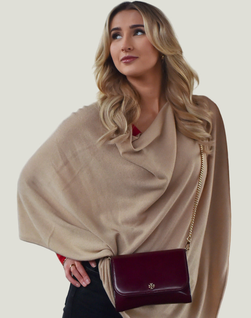 Woman wearing a beige poncho and holding a maroon handbag, standing indoors.