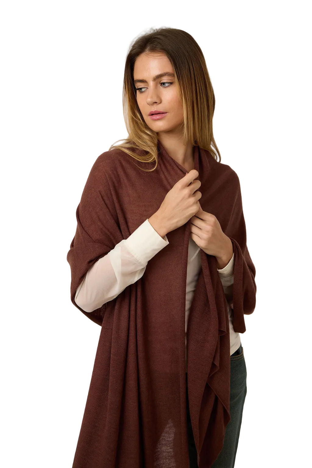 A woman modeling a draped, open-front shawl or wrap in a deep rust-brown color, worn over a white long-sleeve top and dark green pants, against a plain light gray studio background.