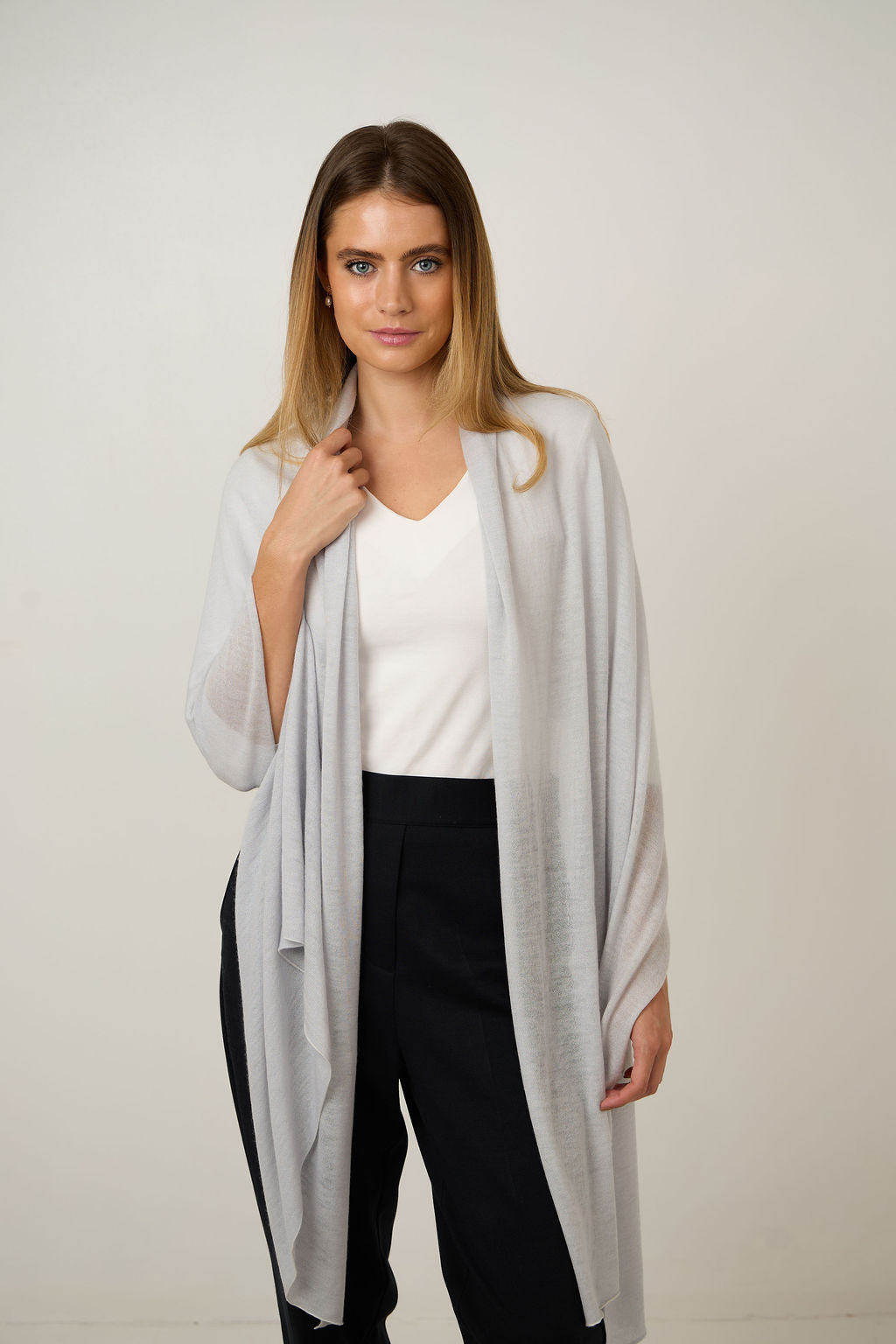 A woman modeling a light gray open-front cardigan over a white V-neck top and black pants, standing against a plain light gray studio background.