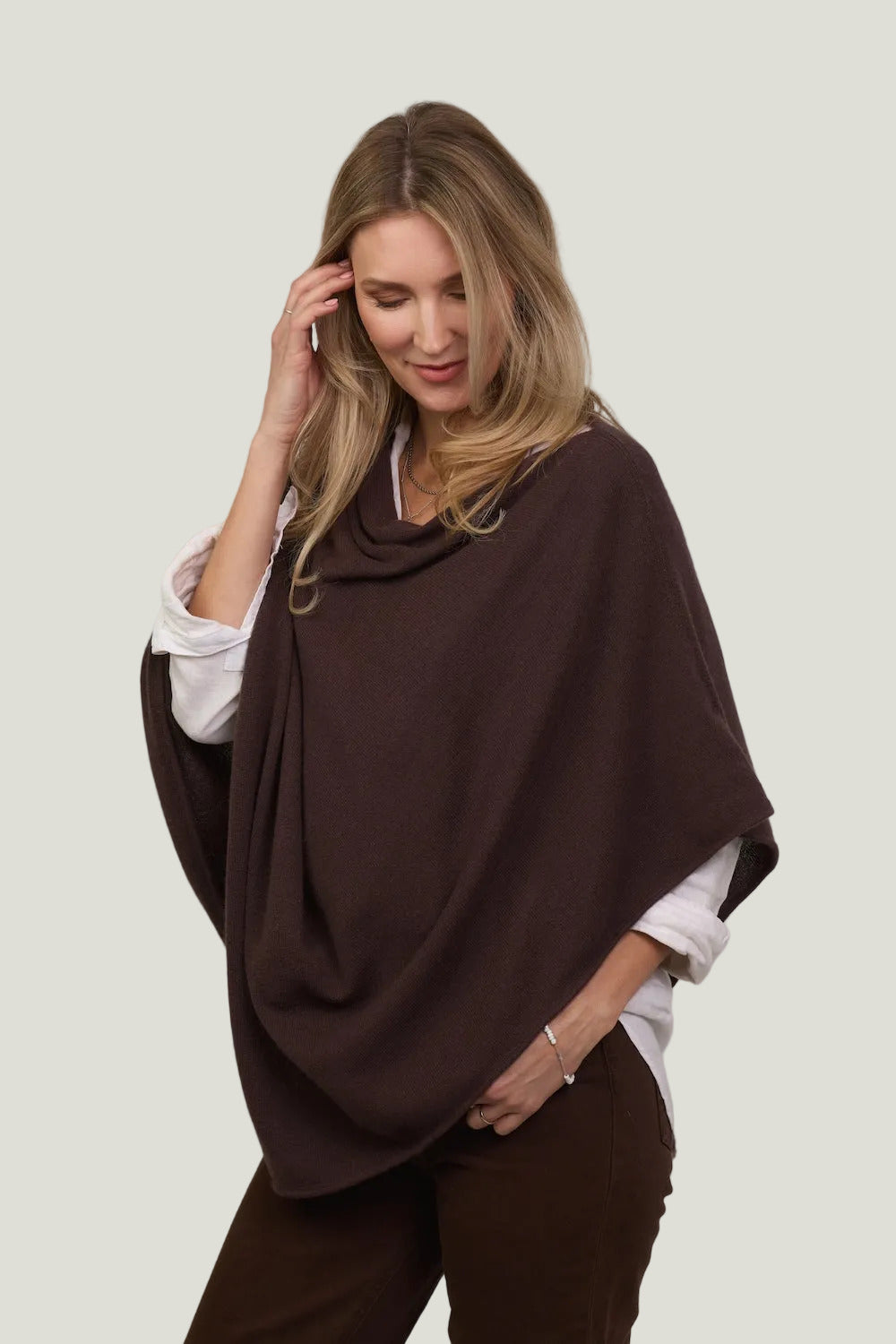 Woman wearing a brown poncho over a white shirt and brown pants on a plain background